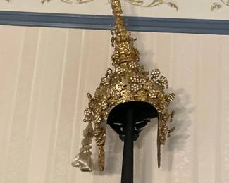Ornate Gold Thai Headdress — Eye-catching ornate gold-tone Thai ceremonial headdress (chada) with tall pointed finial and floral filigree ornamentation. The piece features dangling decorative elements and what appear to be silk or satin ribbon tassels attached at the sides, mounted on a display stand in the photo. Overall measurements are estimates due to photo-only inspection; it's a decorative costume or ceremonial piece appropriate for display rather than everyday wear. Condition appears good with expected surface wear and some tarnish or loss to gold finish on close inspection; tassels show light creasing from storage.