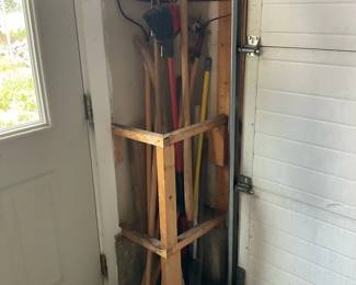 Wooden Garage Tool Corner Organizer — Simple handmade wooden corner organizer built to store rakes, shovels and long-handled garden tools. Constructed from 2x lumber forming two stacked square frames secured to vertical supports; holds multiple tools upright and fits tight in a garage corner by the door. Overall compact footprint makes it easy to install or move; good for utility garages, sheds, or mudrooms and keeps tools organized and off the floor. Condition is functional with surface wear, paint marks, and typical shop/garage dirt but structurally sound; buyer should expect cosmetic imperfections and may wish to sand or refinish.