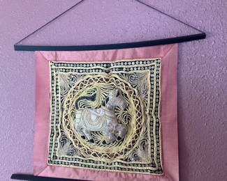 Embroidered Elephant Wall Hanging — Decorative embroidered textile wall hanging featuring a central elephant motif with gold thread, sequins and bead accents on a pink fabric border. The piece is mounted between two slim black wooden hanging bars with a cord for wall display; workmanship appears hand-stitched with metallic embroidery outlining the circular design. Good overall condition with minor surface dust and slight puckering along the fabric corners consistent with display use. Measures approximately 26 inches tall by 16 inches wide; lightweight and ready to hang.