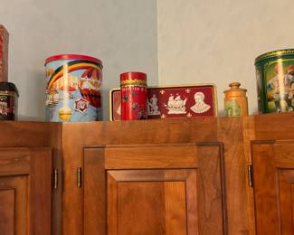 Decorative Metal Tea Tin — A decorative round metal tea tin with colorful printed graphics and a removable lid; pictured among several assorted collectible tins. The tin appears to be mid-size, painted with bright scenes and gold-tone trim visible on some pieces; ideal for storage or display in a kitchen or pantry. Condition is good for display with light surface wear and minor scuffs consistent with shelf use; lid appears to fit. Measurements are approximate.