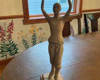 Standing Female Sculpture on Plinth — Tall figurative sculpture of a female dancer or athlete posed with both arms raised, cast in a light-colored material and mounted on a two-tier wooden plinth. The piece shows carved detailing in the clothing and hair, with a naturalistic pose and smooth surface finish. Overall size is moderate and suitable for tabletop or mantel display; estimated measurements provided for hauling and placement. Good estate-sale condition with light surface wear and a few small scuffs on the plinth consistent with age and handling.