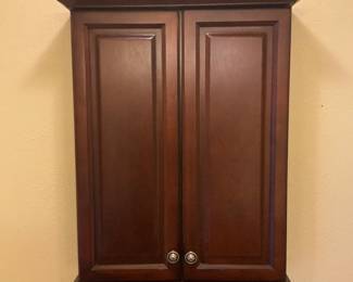 Wall Mounted Cherry Medicine Cabinet — Solid wood wall-mounted medicine cabinet in a warm cherry finish with a molded top cornice, raised-panel double doors and a small shelf at the base. The cabinet shows moderate, even surface wear consistent with normal use; doors close and hardware (two round metal knobs) appears intact. Practical bathroom or hallway storage piece with classic styling and a shallow footprint; exact interior shelving not visible in photos. Measures approximately 36 inches tall, 18 inches wide and 6 inches deep.