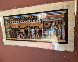 gamal Egyptian Papyrus Print — Hand-signed "gamal" papyrus print depicting a colorful Egyptian funerary scene with hieroglyphs and figures. The print is mounted under clear plastic with scalloped papyrus edges visible; colors remain bright though the papyrus shows age toning and minor edge loss. Signed lower left, it is a decorative reproduction on papyrus typical of tourist or decorative Egyptian-style art. Framed/mounted as shown; condition good with wear to papyrus edges and light surface reflections from the protective covering.