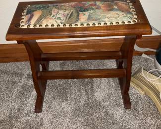 Needlepoint Piano Bench — Small wooden piano or stool bench with a tapestry-style needlepoint upholstered top and brass upholstery tacks. The warm medium-brown wood frame has simple carved stretcher supports and shows light surface wear and a few small scuffs consistent with normal use. The padded top features a multicolor floral/fruit tapestry panel bordered by brass tacks; cushion appears intact though fabric shows light age and slight fading. Compact footprint makes it suitable as piano bench, vanity stool, or entryway seat.
