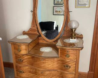 Carved Walnut Vanity Dresser with Mirror — Attractive carved walnut vanity dresser with attached swivel mirror and serpentine front drawers. The piece features figured walnut veneer, carved crest and scroll details, original brass drawer pulls, glass top protectors on the dressing surface, and cabriole legs with brass casters. Solid construction and warm honey finish show light surface wear and small scuffs consistent with normal use but no major structural damage visible. Overall a well-preserved early 20th-century–style bedroom dressing table that measures as noted below and would make a strong focal piece in a traditional bedroom.