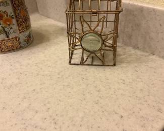 Sun Face Metal Votive Holder — Decorative metal votive holder featuring a sun-face medallion set into a gold-tone wire cage with a small glass cup for a candle. The openwork geometric cage and looped top make it easy to hang or display on a shelf; the glass insert appears intact. Shows light surface wear and patina consistent with age and use but no obvious bends or breaks. Compact decorative piece that works with tealights or small votive candles.