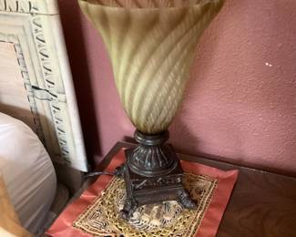 Carved Metal Table Lamp With Glass Shade — Heavy carved metal table lamp with a decorative stepped base and a large swirled amber/olive ribbed glass shade. The metal base shows ornate acanthus-style carving and claw feet with a dark patina; the glass shade has a fluted, diagonal rib pattern and scalloped rim. Works as an accent lamp for a living room or bedroom; lamp appears complete though wiring and bulb function not verified. Overall good solid condition with expected surface wear and patina on the base and minor surface scuffs to the glass.