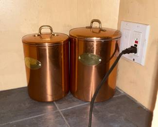 Copper Kitchen Canisters Set of 2 — Pair of cylindrical copper kitchen canisters with lift-off lids and brass-tone loop handles; each has an oval brass label plate on the front. Warm copper finish shows light surface scratches, small dents and some patina consistent with age and use; lids fit but have minor edge wear. Sturdy metal construction makes them useful for dry storage or decorative countertop display; larger canister about twice the diameter of the smaller one.