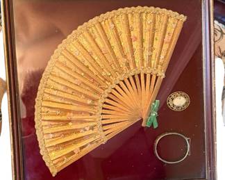 antique framed folding hand fan mounted against a rich red velvet or fabric background in a wooden shadow box, accompanied by a small cameo brooch and a silver-toned hinged bangle bracelet