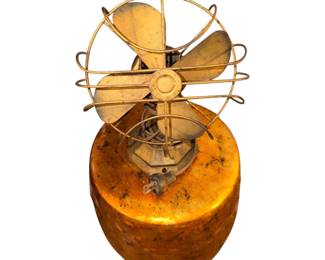 vintage small metal desk fan with a distressed gold finish, four blades, and a circular protective wire cage mounted on a heavy, rounded base.