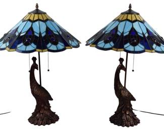 194 - Pr Stained Glass Peacock Table Lamps 27" (Description): A pair of striking stained glass table lamps featuring sculptural peacock bases in a bronze-toned finish. The Tiffany-style shades display an elegant peacock feather motif in shades of blue, cobalt, and cream with decorative glass cabochons. Each lamp features dual pull chain switches. Measures 27 inches tall. Electric/electronic/mechanical item - No guarantee all components, untested.