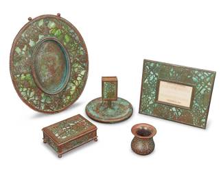 1196 Five Tiffany Studios bronze and Favrile glass 'Grape Leaf' desk items Early 20th century Verdigris patinated, comprising an oval easel-back picture frame, an easel-back calendar holder with incomplete set of paper cards, a matchstick holder, a lidded box, and a bronze brush pot Each impressed: TIFFANY STUDIOS / NEW YORK; match holder numbered: 958; smaller frame impressed with indistinct number 5 pieces Oval aperture 5 5/8 x 3 3/4 in.; rectangular aperture 2 1/4 x 3 3/8 in.; height of box 1 7/8 in., width 4 3/8 in., depth 3 1/8 in.; height of match holder 4 in., diameter 5 1/4 in. Estimate: $800 - $1,200