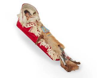 1128 A contemporary Cree beaded pictorial cradle sculpture Attributed to Jack Little Boy (20th/21st century, Canadian Cree) Entitled "Little Chipmunk Face Up Close," circa 1985 The long red velvet cradle with a detailed beaded child's portrait doll face to interior, wrapped in fur, and with hide appliqué fringing to cradle, a small fur pelt to top, and two elaborate beaded pictorial plaques attached to the pelt, one a fringed triangle, and one a mounted warrior, with hide-wrapped handle and hanging loop Unmarked Height 2 ft 6 1/2 in., width 10 1/2 in., depth 9 1/2 in.; pelt tail below cradle 9 in. Estimate: $400 - $600