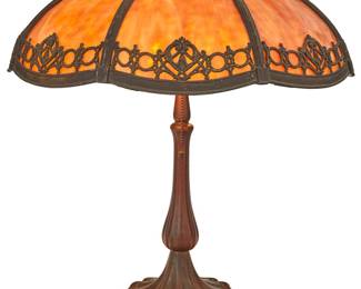 1263 An American mottled glass and metal overlay table lamp Early 20th century, possibly Handel Amber slag glass shade with decorative metal overlay design, raised on a patinated metal four-lobed base issuing two lights with acorn pulls, electrified Unmarked Height overall 21 1/2 in.; height of shade 6 1/4 in., diameter 18 3/4 in. Estimate: $800 - $1,200