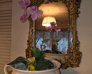 Vintage Chamber Pott with potted silk Orchid inside for decoration;  Baroque/Rococo style Ornate Gold Gilded Wall Mirror embellished with flowing scrolls, stylized foliage and decorative crest