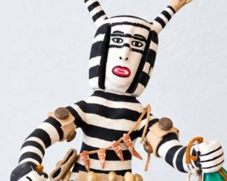 Black-and-white striped Kachina-style figure with horned headpiece, shell accents, leather details and handheld accessories.