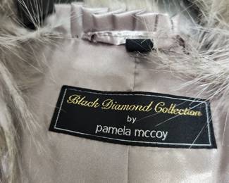 full-length faux fur coat from the Pamela McCoy Black Diamond Collection. This specific line is highly regarded for its premium, heavy-weight replica furs designed to mimic natural textures like coyote, wolf, or fox. The garment features a long-pile, shaggy finish with multi-tonal gray, cream, and brown striations, giving it a realistic animal-print aesthetic.