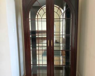 Tall mahogany finish display cabinet featuring a dramatic broken-scroll |bonnet pediment with center urn finial |twin full-length glass doors, decorative arched wood mullions, brass-tone hardware and carved detailing at the base | Inside 3 glass shelves plus interior lighting