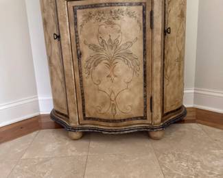 Decorative hand-painted bombe-style cabinet in an antique cream/gold finish with dark bronze accents. Features a shaped serpentine top and base, ornate floral and scroll detailing, one upper drawer with decorative metal pull, a center door and two curved side storage doors with interior shelving.