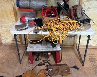 Craftsman Benchtop Table Saw with Circular Saw and Accessories This lot centers on a Craftsman benchtop table saw on a folding metal stand with the "CRAFTSMAN" nameplate clearly visible on the front rail. Included alongside are a corded circular saw, a heavy-duty yellow outdoor extension cord, and a leather carpenter's tool belt with multiple pouches. The table saw shows honest workshop wear with surface dust and staining on the deck, and the folding stand makes it portable between jobsite and shop. Working condition not confirmed on all items — sold as found.