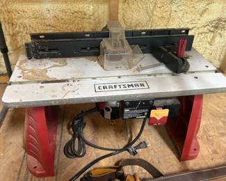 This Craftsman router table sits on sturdy red molded legs and features an adjustable split fence, clear blade guard, miter slot, and a safety power switch with easy-reach paddle. The "CRAFTSMAN" branding is clearly marked on the aluminum table surface. Craftsman router tables are a reliable choice for edge profiling, dado cuts, and decorative woodworking, and this one shows moderate workshop use with surface staining on the tabletop. Working condition not confirmed — sold as found.