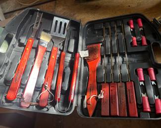 BBQ Grilling Tool Set in Molded Hard Case This complete barbecue grilling tool set comes in a black molded hard case and includes stainless steel utensils with red wood-look handles: a slotted spatula, locking tongs, carving fork, basting brush, grill cleaning brush, four steak knives, corn cob holders, and metal skewers — all fitted into die-cut slots. A ready-to-gift BBQ tool kit for grilling season, tailgating, or setting up a backyard cookout station. The tools and case appear in lightly used condition with all original pieces present.
