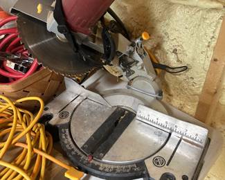 This compound miter saw features a large blade with carbide-tipped teeth, an adjustable miter gauge with marked detents, and an attached dust collection bag for cleaner cuts. The saw shows honest workshop use throughout with surface dust and wear on the base plate, and the yellow/orange handle accents suggest it may be a DeWalt or similar brand — confirm the nameplate in person. A solid compound miter saw for woodworking, framing, or trim projects. Working condition not confirmed — sold as found.