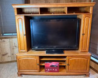 Oak Entertainment Center with Hitachi TV and Sony DVD Player This honey-oak entertainment center features arched glass-panel cabinet doors on the upper section, open shelving in the center for a TV, and lower storage compartments. A Hitachi flat-screen TV sits in the center bay, and a Sony DVP-SR510H DVD player is included — appears to be in or near its original packaging. Oak entertainment centers remain a functional living room storage solution with room for media equipment, books, and display items. Working condition not confirmed on electronics — sold as found.