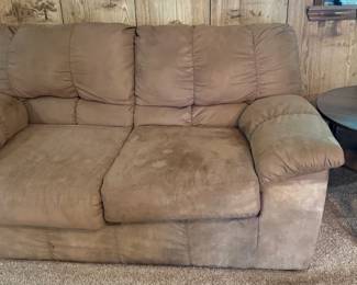 Brown Microsuede Loveseat with Dark Wood End Table This brown microsuede loveseat features padded cushion seats and pillow backs in a warm chocolate tone. The cushions show visible staining and wear on the seat surfaces consistent with regular household use — condition should be evaluated in person. Included is a round dark wood end table with a turned pedestal leg and a lower display shelf. The loveseat and table together make a functional living room or den seating pair, and the loveseat frame appears solid beneath the upholstery.