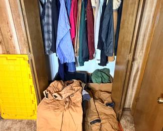 Men's Coat Closet Lot — Jackets, Outerwear, and Field Gear This men's coat closet lot includes a full hanging rod of jackets and outerwear: visible pieces include a plaid flannel shirt jacket, a blue windbreaker, a dark green field coat, and several other lined and layered coats in assorted sizes. On the closet floor sit a pair of tan duck canvas duffel bags or rolled insulated coveralls and a yellow folding utility stool. A practical men's outerwear lot covering work jackets, hunting layers, and cold-weather coats in one grouping — confirm sizes in person.