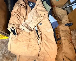This lot features a pair of Walls brand insulated coveralls in classic tan duck canvas with a corduroy-trimmed collar, front zipper, and ankle zippers for easy on-and-off over boots. The coveralls show moderate use with some soiling consistent with outdoor work. Additional outerwear items visible in the lot include a green jacket and other layering pieces in storage bins. Walls insulated coveralls are a cold-weather workwear staple for hunting, farming, and outdoor jobs.