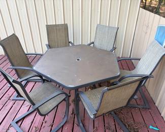 Hexagonal Patio Dining Set — Table with 6 Sling Chairs This outdoor patio dining set includes a hexagonal metal-top table with umbrella hole and six C-spring sling-back dining chairs in coordinating earth-tone fabrics. The dark bronze-finished steel frames are sturdy and weather-resistant, and the high-back sling seats provide comfortable spring-action motion. The set shows moderate outdoor use with surface weathering on the tabletop. A complete 7-piece patio dining set ready for a deck, porch, or backyard entertaining space.