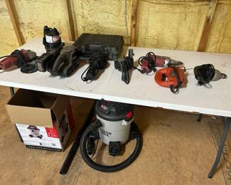 This lot includes an assortment of corded power tools laid out on a folding table: a belt sander, a finishing sander, what appears to be a rotary tool or die grinder, a compact angle grinder, an orange jigsaw, and a hard-sided tool case with contents. Below the table sits a Porter-Cable wet/dry shop vacuum with hose and its original box. A well-rounded corded power tool collection for a home workshop or jobsite, covering sanding, cutting, and cleanup in one lot. Working condition not confirmed on all items — sold as found.