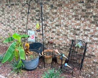 This garden decor lot includes two wrought iron shepherd's hook plant hangers, a "Love Grows Here" decorative garden flag, a blown glass hummingbird feeder, a large gray resin planter pot, a terra cotta pot, a small black metal three-tier plant stand, a flamingo yard ornament, a turtle garden figurine, and several colorful ceramic mushroom garden stakes. A ready-made garden accent collection that covers hanging, potting, and decorative display in one lot.