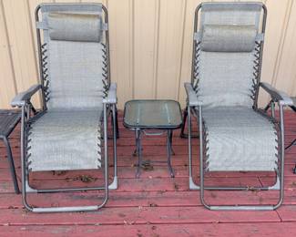 This three-piece patio set includes two folding zero gravity lounge chairs in gray textured sling fabric with padded headrest pillows, plus a small folding glass-top side table. Zero gravity reclining chairs are popular for patio relaxation, poolside lounging, and camping, and this set's neutral gray color coordinates with most outdoor spaces. The chairs show light use with some minor discoloration on the headrests, and the side table glass is intact.