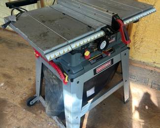 Craftsman 10-Inch Table Saw on Floor Stand This Craftsman 10-inch table saw sits on a sturdy open-leg metal floor stand with the "CRAFTSMAN" nameplate and "10 in" blade size clearly marked on the upper blade guard housing. The saw features a cast-iron table surface with extension wings, a rip fence, miter gauge slot, and a red-and-yellow safety paddle switch for quick shutoff. The blade appears to still have its factory protective wrap in place, and the overall unit shows moderate workshop dust and use. A reliable 10-inch table saw for ripping plywood, crosscutting lumber, and general shop woodworking. Working condition not confirmed — sold as found.