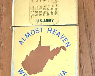 a vintage flip-top windproof pocket lighter featuring a "July 1977" calendar graphic, an insignia for the U.S. Army, and a souvenir map print of West Virginia captioned with the iconic John Denver lyric, "Almost Heaven".
