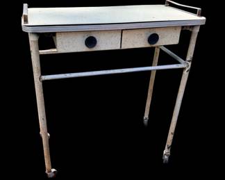 vintage, off-white enameled metal medical or salon utility table, likely from the mid-20th century, featuring two small drawers with dark circular pulls, a lower tubular stretcher bar, and caster wheels