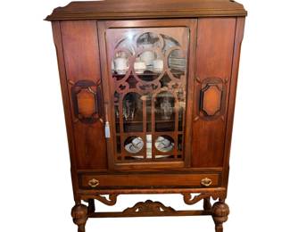 This is an antique 1920s Jacobean Revival style walnut china cabinet featuring a single glazed door with fretwork, a lower drawer, and bulbous cup-and-cover turned legs.