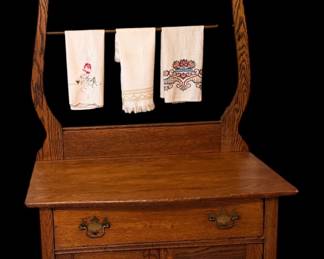 This an early 1900s antique solid oak washstand commode featuring a high-back harp, a beveled mirror, and an integrated towel bar.
