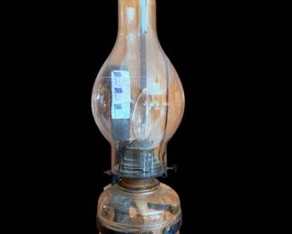 rustic, vintage-style glass oil lamp or hurricane lamp with a clear glass font and chimney, featuring a dark metal holder and a handle or base strap.