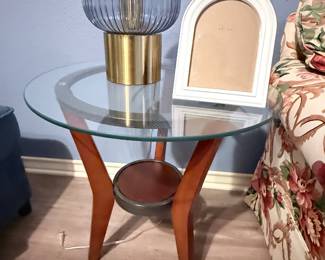 **Glass-Top Accent Table** – Round glass-top accent table featuring a wood base with a lower display shelf. Shown with a contemporary table lamp. A versatile piece for a living room, bedroom, or sitting area. Items sold individually unless noted.