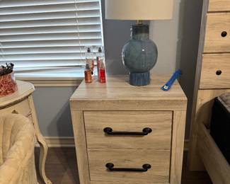 Two-drawer nightstand featuring a light wood finish and black hardware, paired with a blue ceramic table lamp. Shown with decorative perfume bottles and small tabletop accessories. Items sold individually unless noted.