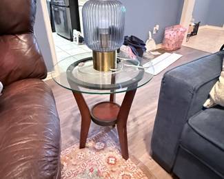 **Glass-Top Accent Table** – Round glass-top accent table featuring a wood base with a lower display shelf. Shown with a contemporary table lamp. A versatile piece for a living room, bedroom, or sitting area. Items sold individually unless noted.