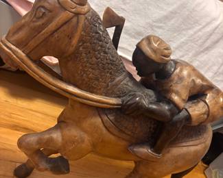 Hand-carved wooden sculpture depicts a jockey in a racing pose atop a horse. This piece is crafted from solid wood with a two-tone finish, featuring dark stained accents on the jockey's skin and the horse's mane and tail. The horse has a textured saddle and bridle details. A wooden axe-like element is integrated behind the jockey's seat.