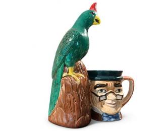 Ceramic figural parrot tiki mug with face handle
Condition: Good
Glossy ceramic novelty mug shaped as a parrot perched on a carved base, with a stylized face integrated into the mug body/handle area. Bright green parrot, red crest, yellow beak/feet, and brown textured base. Likely a tiki/souvenir collectible mug.