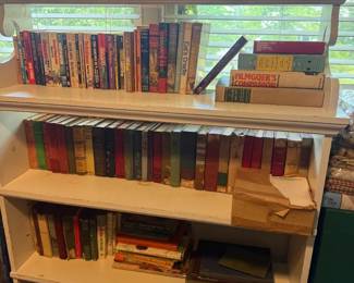 White wooden adjustable-shelf bookcase
Condition: Good
White-painted wooden bookcase with multiple open shelves and visible wear from use. The shelves are filled with assorted paperback and hardcover books; a few small boxes and miscellaneous items are also present. Appears to be a practical storage piece rather than a designer or collectible bookcase.