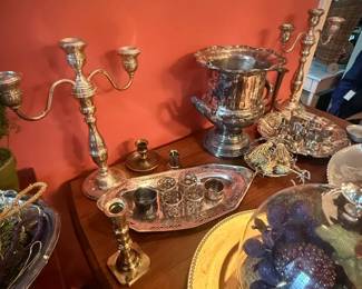 Silverplate decorative tableware lot with candelabra, ice bucket, and tray
Condition: Good
Assorted silver-colored metal table/display pieces on a tabletop, likely silverplate rather than sterling based on appearance. Visible items include several candlesticks/candelabra, a large trophy-form ice bucket or urn, trays, and small serving accessories.
