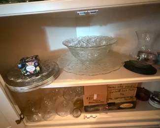 Clear Pressed Glass Pedestal Punch Bowl / Compote Centerpiece
Condition: Good
Large clear glass pedestal bowl with a scalloped rim and ornate pressed/cut pattern, displayed on a cabinet shelf among other glassware. The form could function as a punch bowl, compote, or centerpiece bowl; the photo does not prove a matched set or maker.