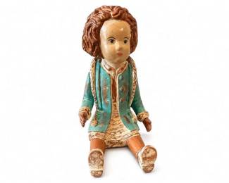 Hand-carved wooden seated folk art doll
Condition: Fair
Seated carved wood doll/figure with painted facial features, curly brown hair, turquoise coat with gold trim, tan skirt/legs, and significant paint loss/wear on the legs, shoes, and edges. The form resembles a folk-art or artist-made wooden doll rather than a mass-produced toy.